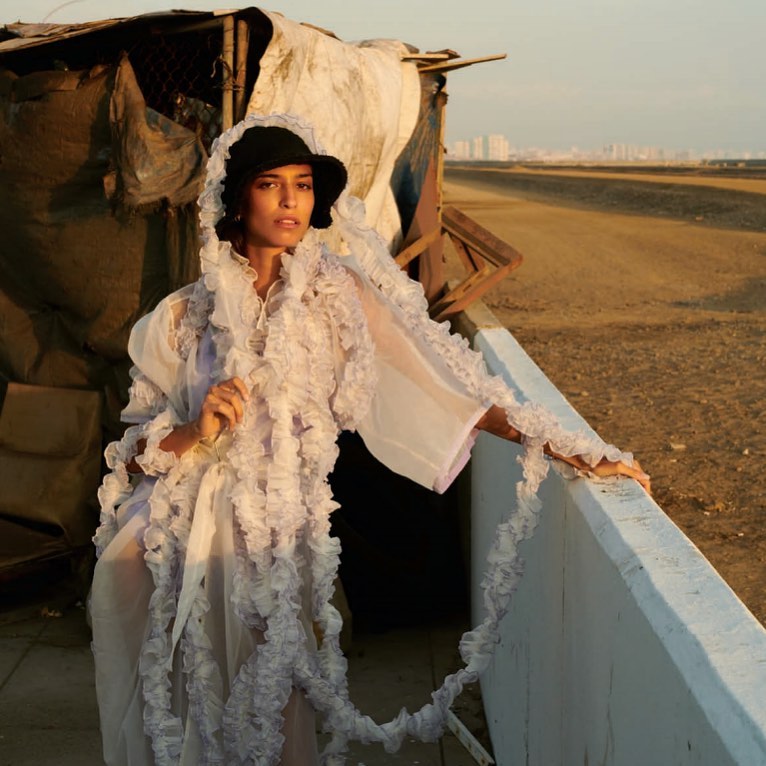 Adult female fashion model wears a ruffled sheer robe and hat, posing outdoors with an urban cityscape in the background.