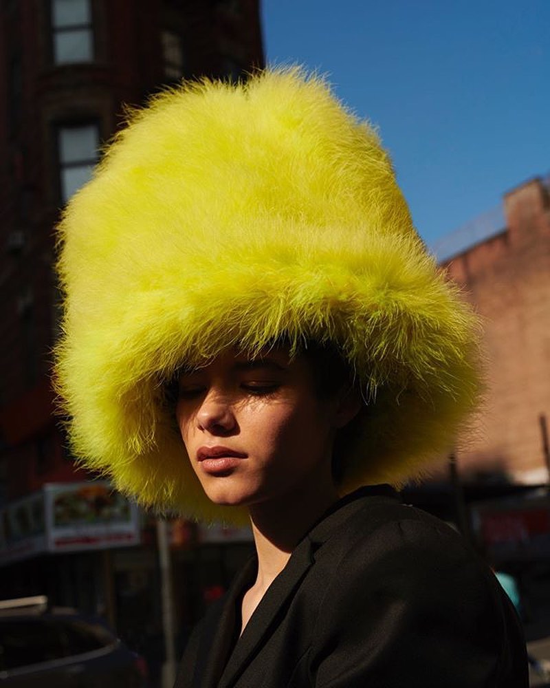 Woman with dark hair wearing a bright yellow fluffy hat and a black jacket, posing outdoors in an urban environment during daylight.
