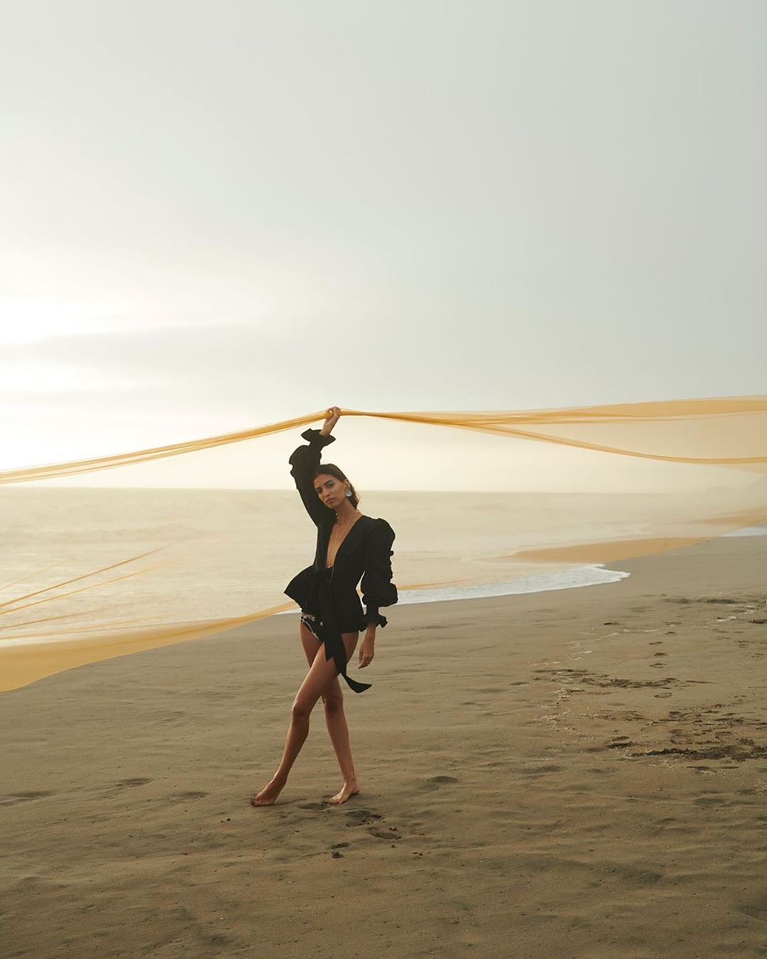 Model in a black blouse holding an orange fabric ribbon on a sandy beach at sunset by Alexander Neumann