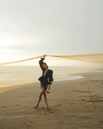 Model in a black blouse holding an orange fabric ribbon on a sandy beach at sunset by Alexander Neumann