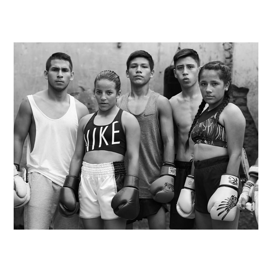 Black and white portrait of five young boxers wearing boxing gloves and sportswear standing in front of a textured wall, looking at the camera.
