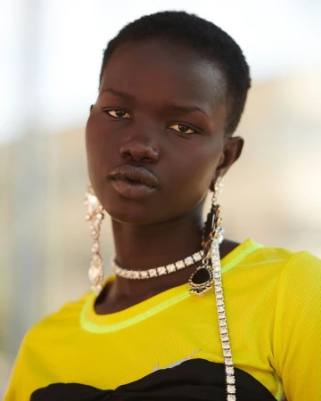 Close-up of an African-descent adult female model with short hair wearing a yellow-and-black top and jewelry, shot indoors.