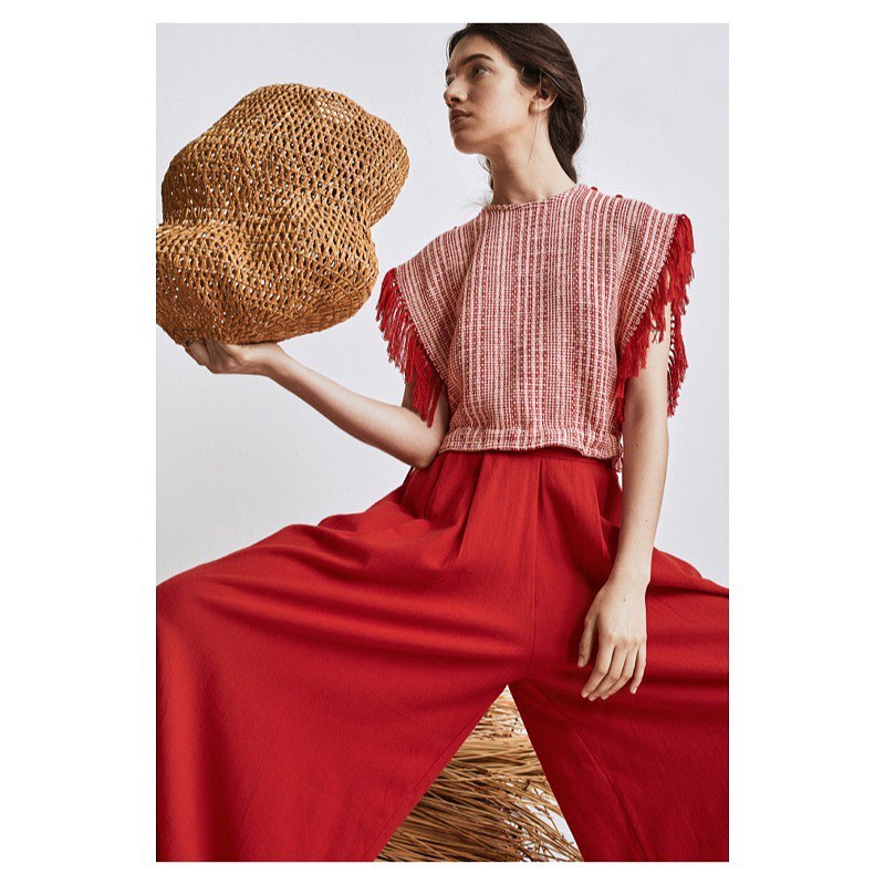 Woman in a red, textured crop top with fringe details and high-waisted, wide-leg red pants posing with a wicker sculpture indoors.