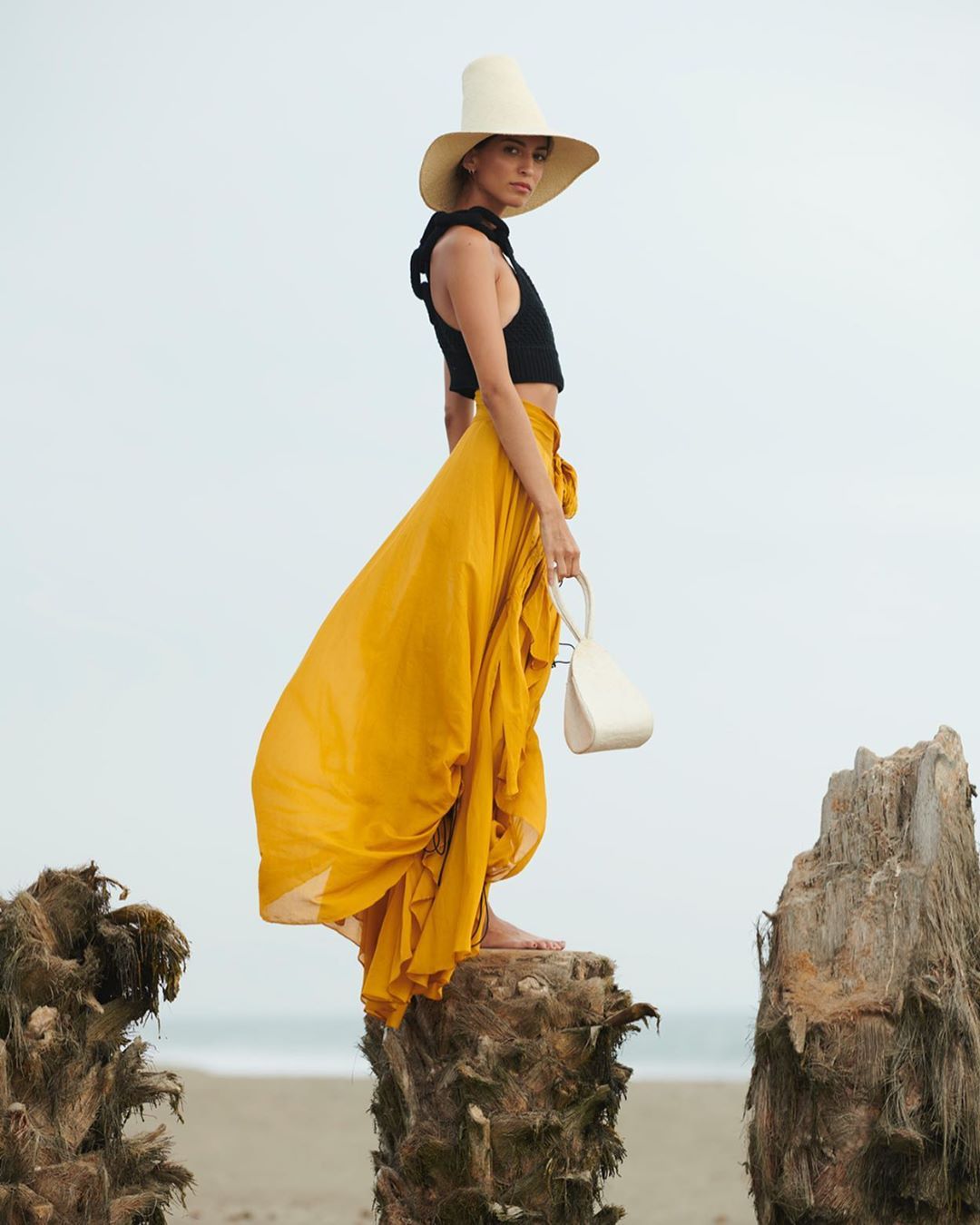 A woman in a black top and yellow skirt stands on a tree stump by the beach with a white hat and purse in an outdoor fashion shot.