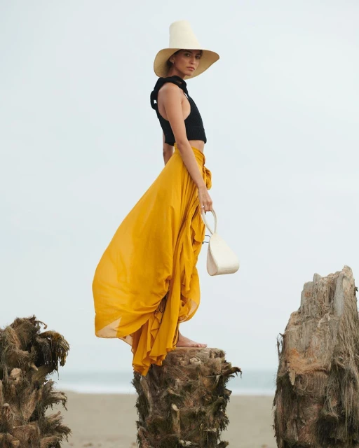 A woman in a black top and yellow skirt stands on a tree stump by the beach with a white hat and purse in an outdoor fashion shot.