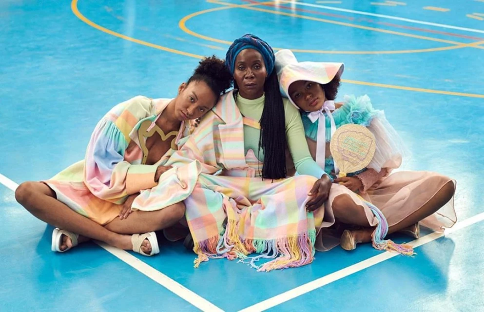 Three Black women in pastel, vintage-inspired outfits sit cross-legged on a bright blue indoor court, radiating style and confidence.