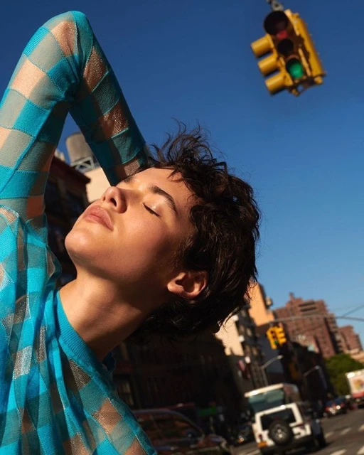 Woman in blue checkered top stands in city street with eyes closed, backlit by the sun against the urban backdrop and blue sky.