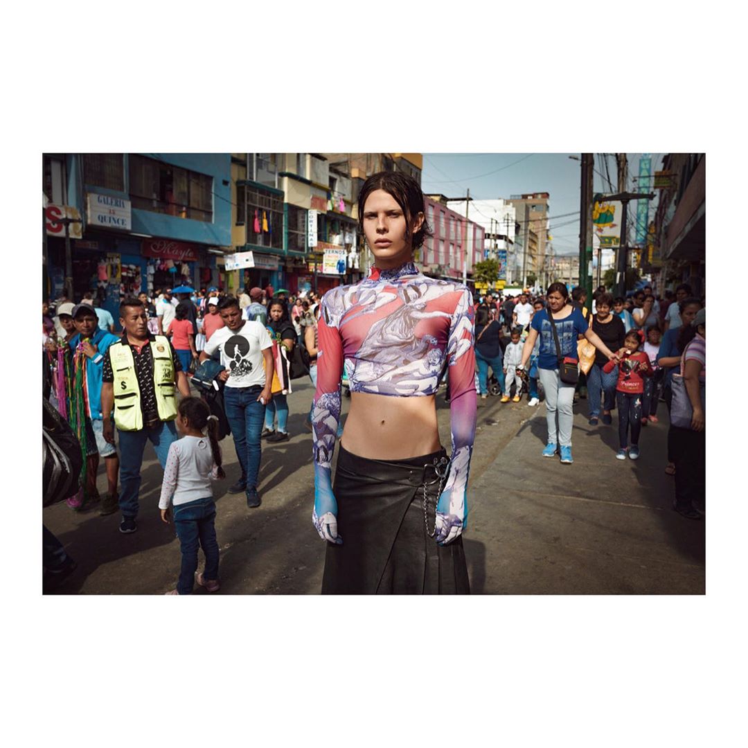 Fashion editorial shot of androgynous model in printed crop top and black skirt standing on a busy street surrounded by pedestrians.