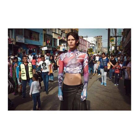 Fashion editorial shot of androgynous model in printed crop top and black skirt standing on a busy street surrounded by pedestrians.