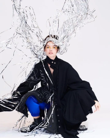 An adult woman in a long black coat with silver tinsel 'hair' poses in a photo studio. She wears blue pants and zebra-striped boots.