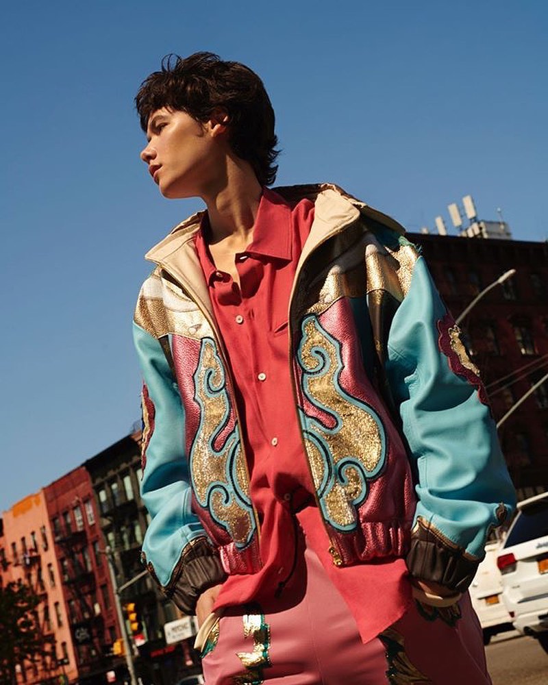 Model in a colorful jacket and pink suit stands on an urban street under a blue sky. Buildings and a vehicle are visible.