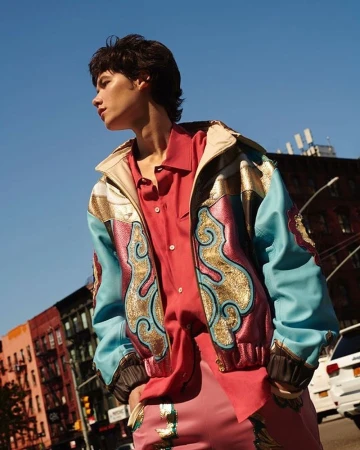 Model in a colorful jacket and pink suit stands on an urban street under a blue sky. Buildings and a vehicle are visible.