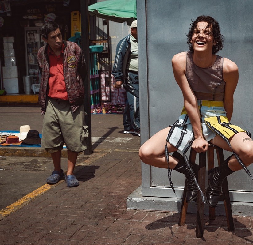 A fashion-forward person sits on a stool on a city street with two other people visible in the background on a sunny day.