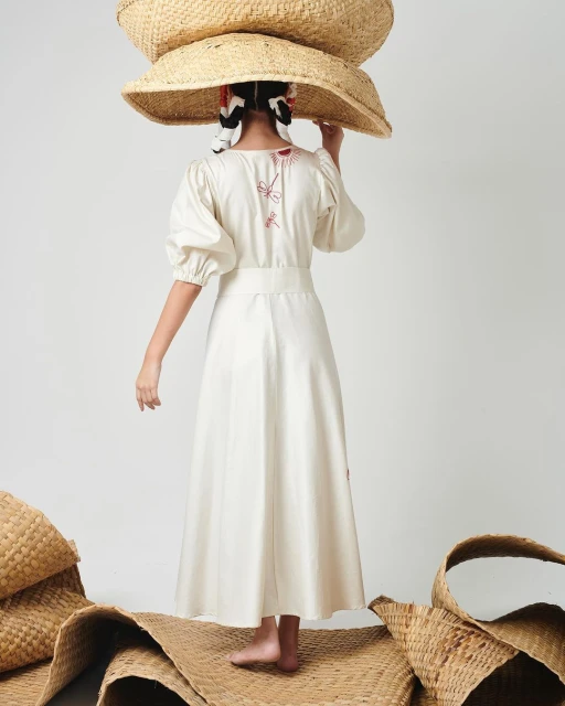 A young girl is seen from the back wearing a white dress and three straw hats stacked on her head. She stands on a woven mat.