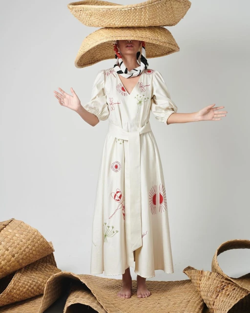 Woman in an embroidered white dress stands on a woven mat with hands raised, wearing two woven hats and a patterned scarf in a white studio.