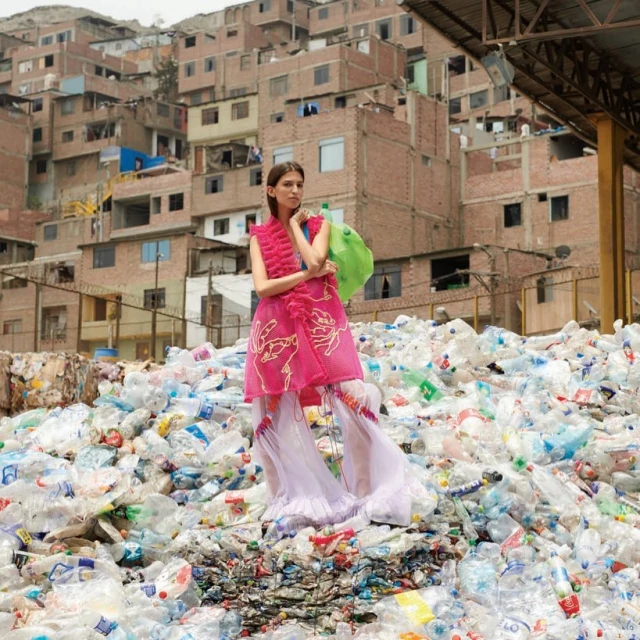 Fashion model in a bright pink dress posing on a heap of plastic bottles with a dense urban backdrop, blending fashion with environmental themes.
