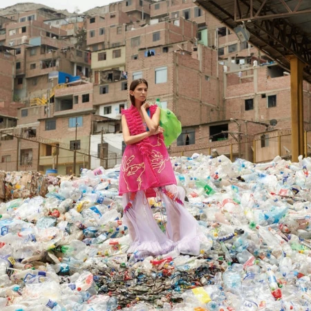 Fashion model in a bright pink dress posing on a heap of plastic bottles with a dense urban backdrop, blending fashion with environmental themes.