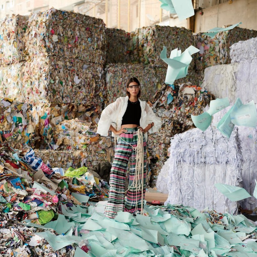 A woman in patterned pants and jacket poses amid towering stacks of recycled paper and scattered paper scraps in a factory or warehouse.