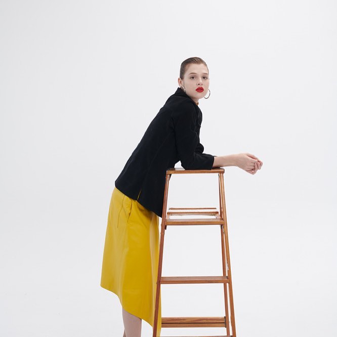 Young woman in a black blazer and yellow skirt leans on a wooden step stool in a bright studio with a neutral expression and red lipstick.