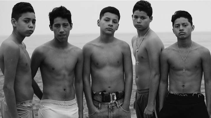 Black and white portrait of five Hispanic teen boys standing on a beach in swim trunks or jeans, and wearing necklaces.