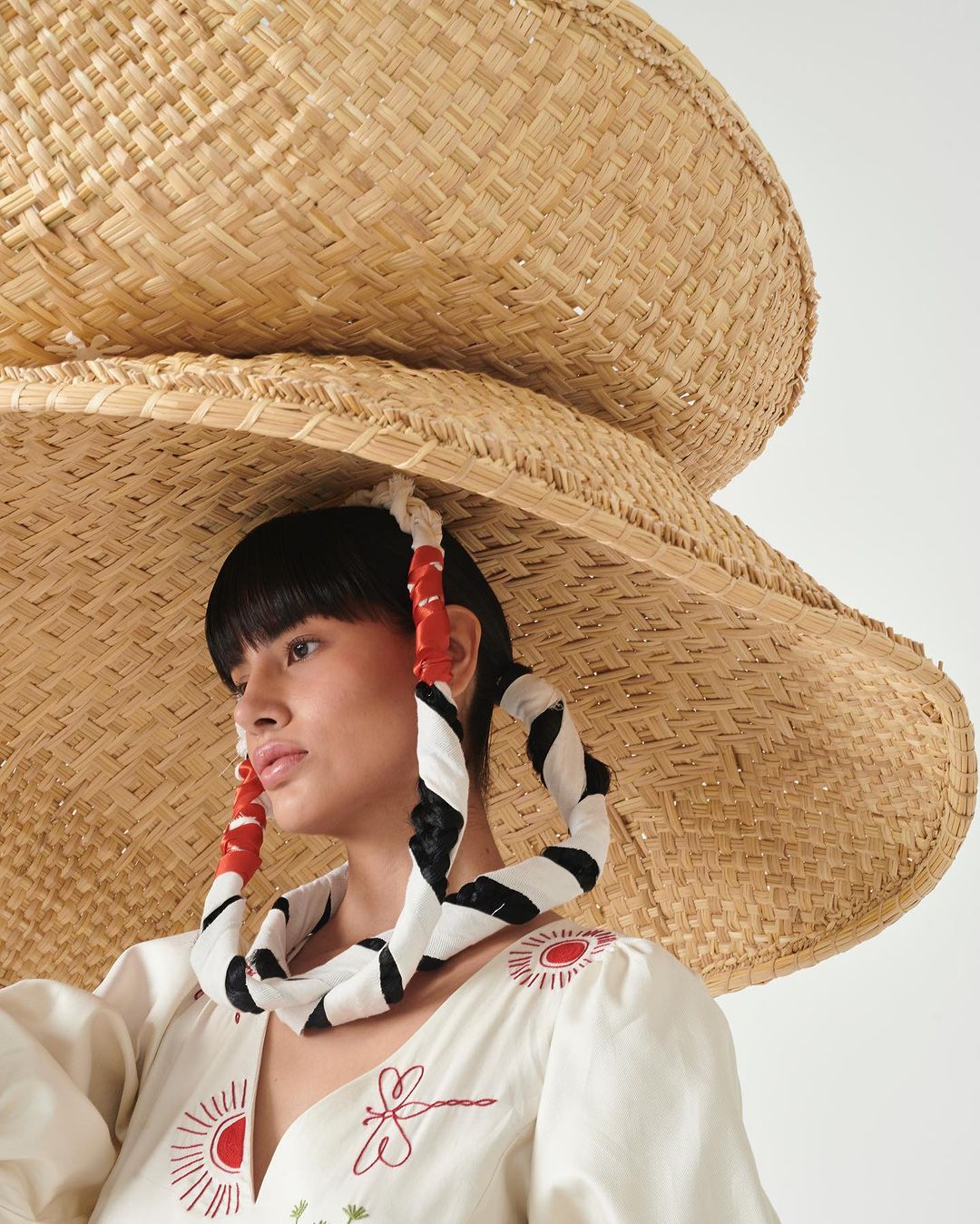 Woman with dramatic straw hat and striped fabric necklace against a white backdrop in a studio setting. Fashion and beauty shot.