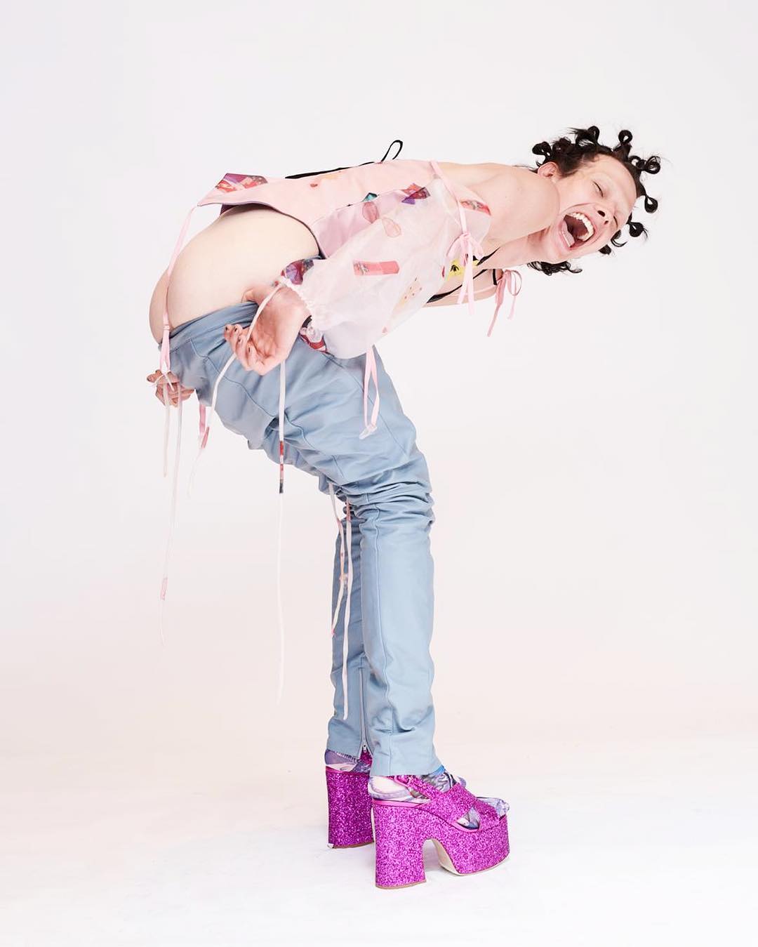A person arches backwards wearing a sheer pink top, blue jeans, and glittery platform shoes against a bright white studio backdrop.