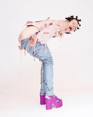 A person arches backwards wearing a sheer pink top, blue jeans, and glittery platform shoes against a bright white studio backdrop.
