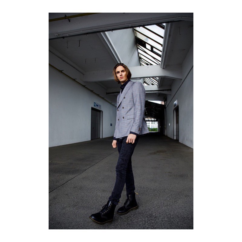 A stylish adult male walking in an industrial-style interior wearing a grey suit jacket, black turtle neck, dark jeans, and black boots.