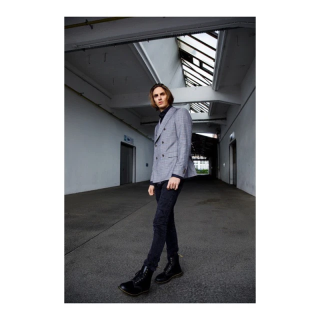 A stylish adult male walking in an industrial-style interior wearing a grey suit jacket, black turtle neck, dark jeans, and black boots.