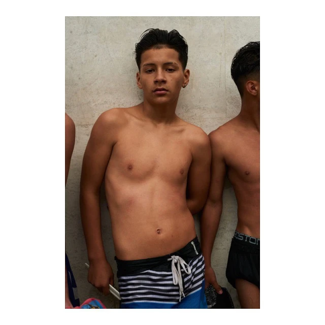 A shirtless young man in swim trunks leans against a concrete wall next to another young man; medium shot, natural lighting.
