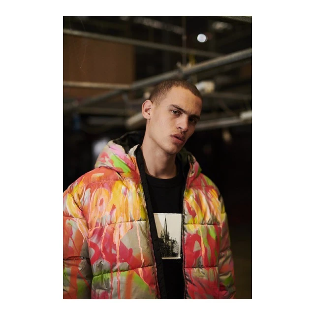 Adult male model wearing a colorful, patterned puffer jacket, black t-shirt with a building graphic, indoors with industrial ceiling.