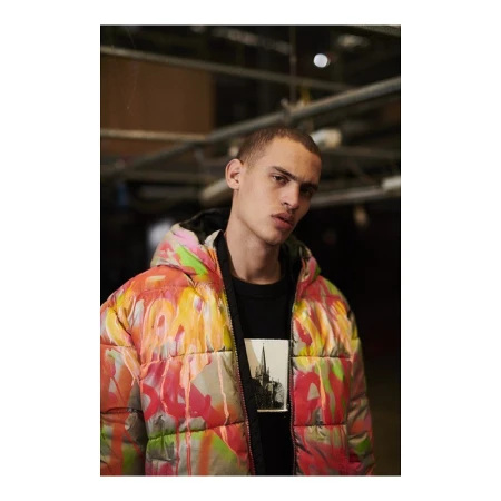 Adult male model wearing a colorful, patterned puffer jacket, black t-shirt with a building graphic, indoors with industrial ceiling.