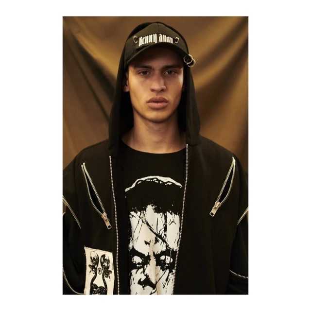 Close-up portrait of a young adult male in a black hooded jacket and ball cap with metal rings, standing against a warm brown backdrop.