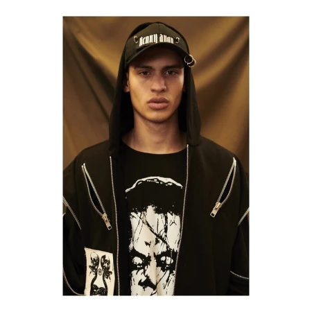 Close-up portrait of a young adult male in a black hooded jacket and ball cap with metal rings, standing against a warm brown backdrop.