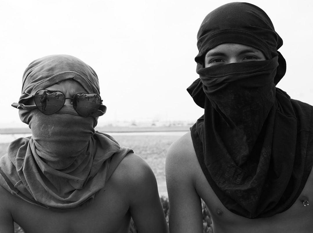 Black-and-white portrait of two young adults with partially obscured faces, scarves, and sunglasses against an outdoor background.