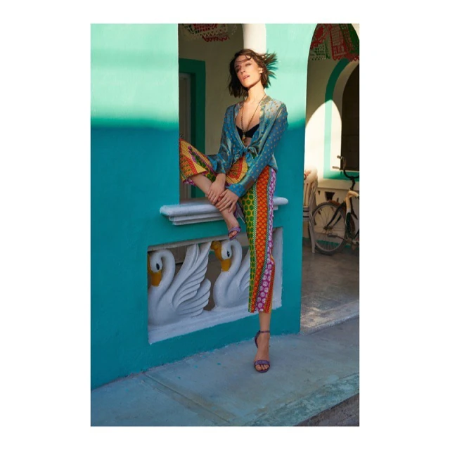 A stylish woman poses in a colorful turquoise building, wearing patterned pants and a silk blouse in soft daylight. She sits by a bicycle.