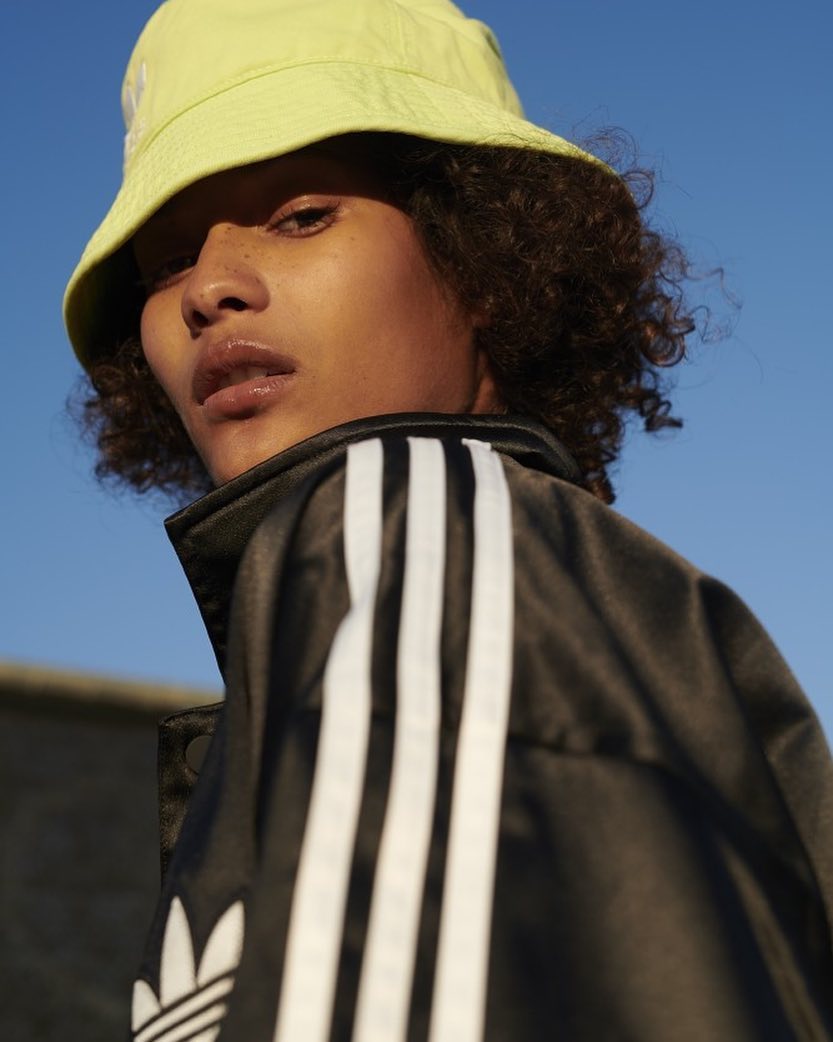 Portrait of an adult female with dark curly hair, wearing a lime green bucket hat and a black Adidas jacket with white stripes against a blue sky.
