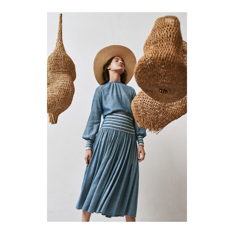 A woman in a blue dress and wide-brimmed hat poses indoors with woven pendant lights, creating a dreamy and fashionable atmosphere.