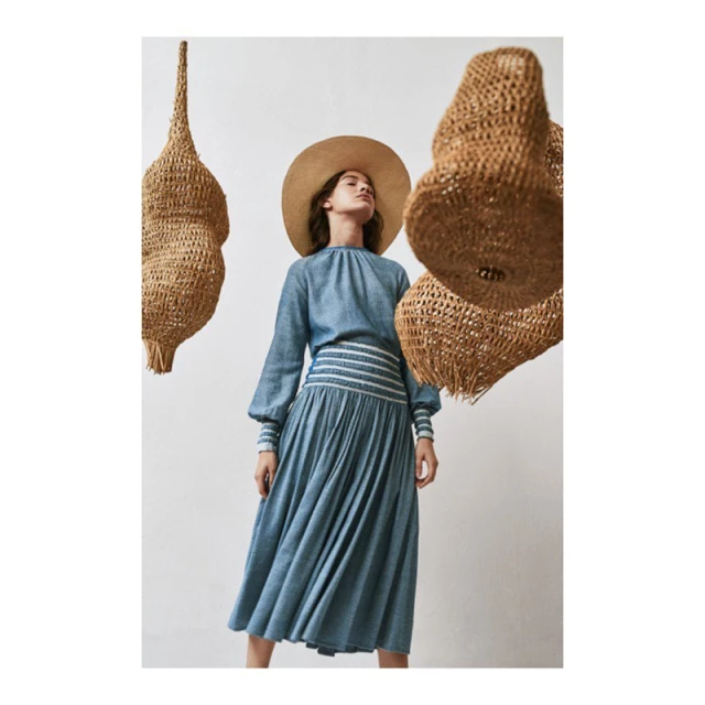 A woman in a blue dress and wide-brimmed hat poses indoors with woven pendant lights, creating a dreamy and fashionable atmosphere.