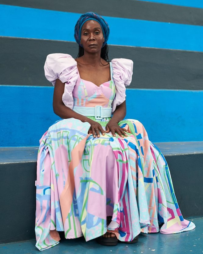 Elegant African-American woman sits on blue steps in a long, colorful dress with puffed sleeves and a blue headwrap, posing gracefully.