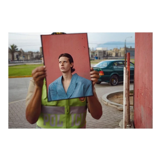 A person in a reflective vest holds up a framed photo of a person in a blue suit on an overcast street with a car and buildings.