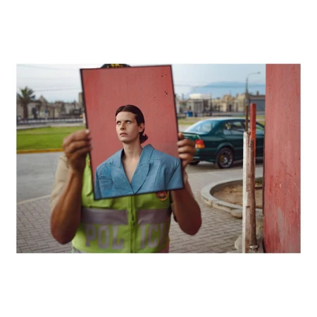 A person in a reflective vest holds up a framed photo of a person in a blue suit on an overcast street with a car and buildings.