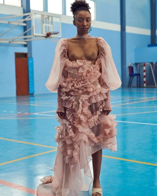 Woman poses in a pink ruffled dress in a gymnasium. Fashion shot with athletic background. Indoor studio-style setting and neutral color palette.