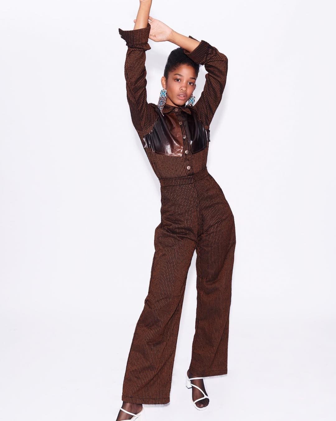 A young adult woman poses in a brown and black leather-accented pantsuit with her arms raised against a plain white backdrop.