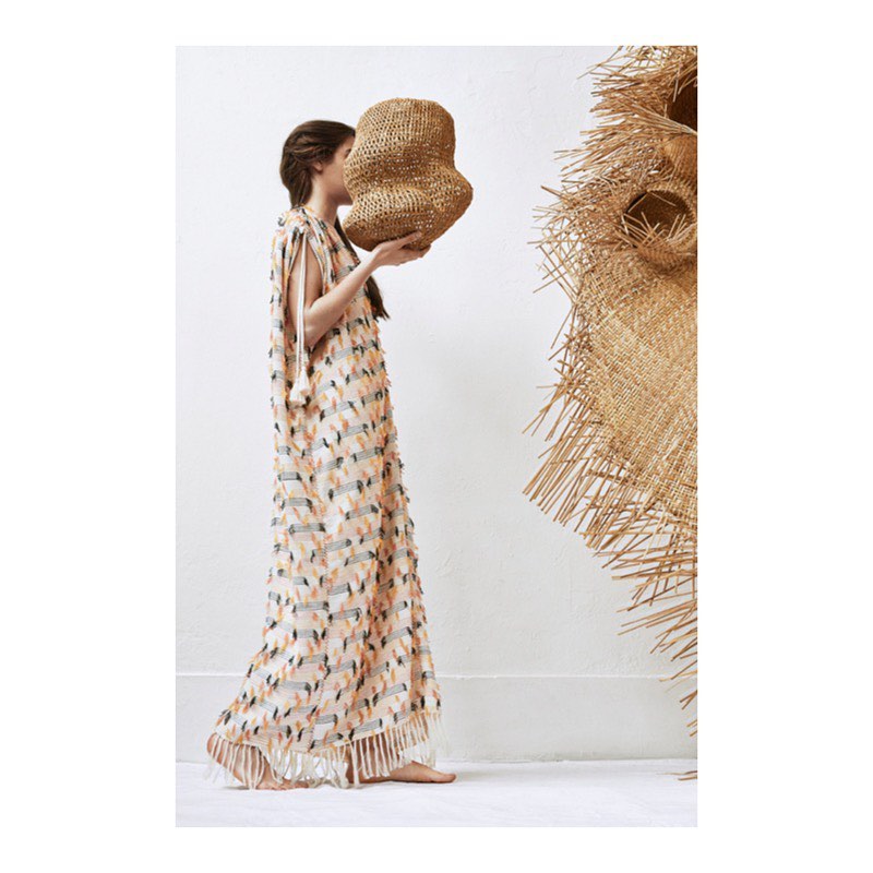 Woman in patterned dress holds woven object in minimalist setting with a large straw decoration; side view, fair-skinned.