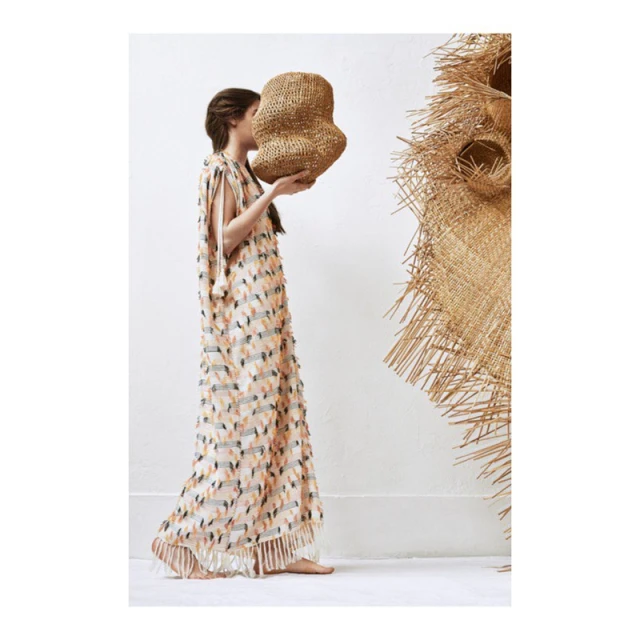 Woman in patterned dress holds woven object in minimalist setting with a large straw decoration; side view, fair-skinned.