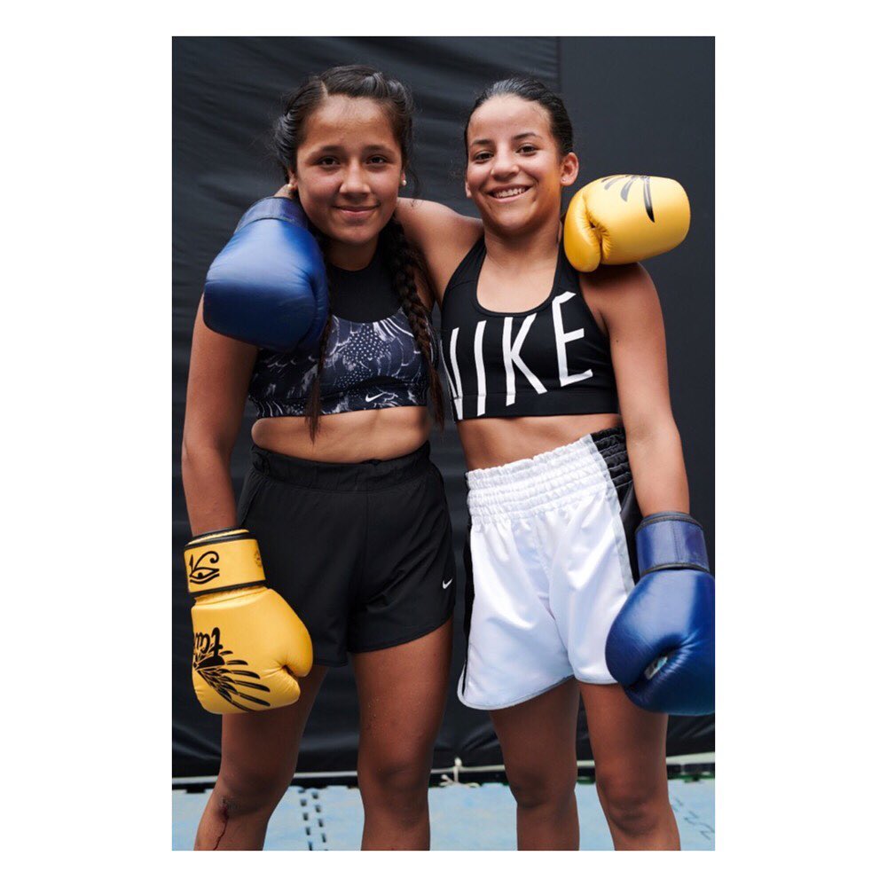 Two young women with boxing gloves posing indoors, wearing athletic tops and shorts, with yellow and blue gloves and braided hair.