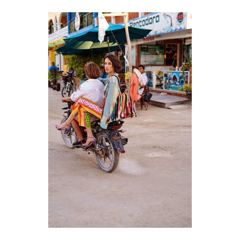 Two women ride a scooter down a sunny street with shops in the background, one looking at the camera, wearing colorful clothing.
