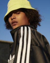 Model wearing a bright yellow bucket hat and a black track jacket with white stripes against a clear blue sky.