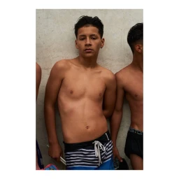 Shirtless teenage male model wearing blue and black striped boardshorts against a neutral wall by Alexander Neumann